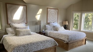 Un dormitorio con dos camas, cada una con estructura de madera y ropa de cama estampada. Hay dos mesitas de noche con lámparas y un ventanal grande que deja entrar luz natural.