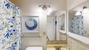 A bathroom with a shower curtain featuring a blue and green floral pattern, a wall-mounted mirror, and a framed abstract art piece.
