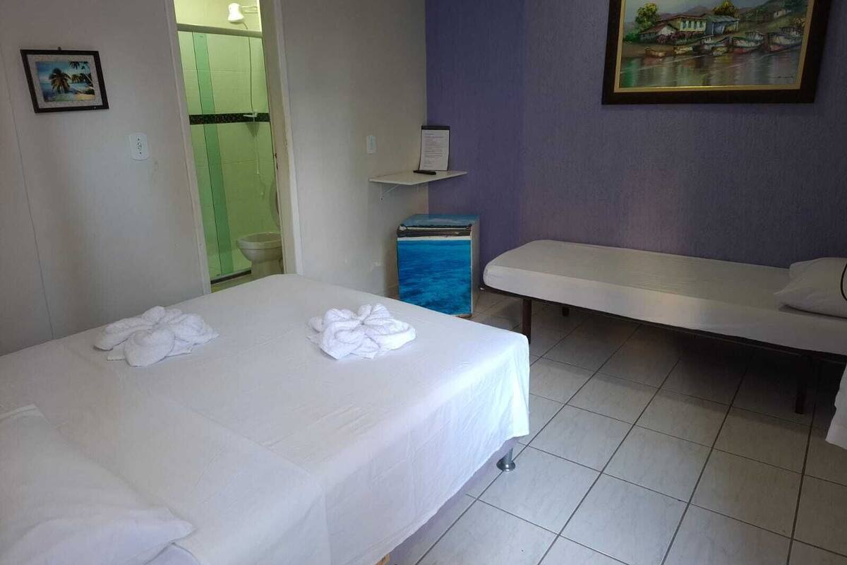 Quarto de hotel com cama, um banquinho, uma pintura e uma pequena estante.