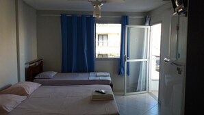 Habitación de hotel con dos camas, ventilador de techo, puerta corrediza de vidrio al balcón y televisor en la pared.
