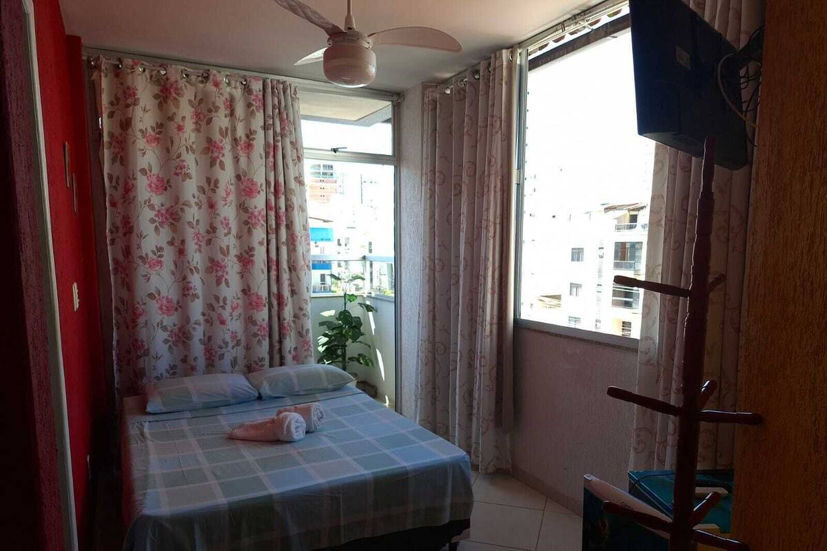 Quarto com cama, cortinas, ventilador de teto e janela com vista para edifícios.