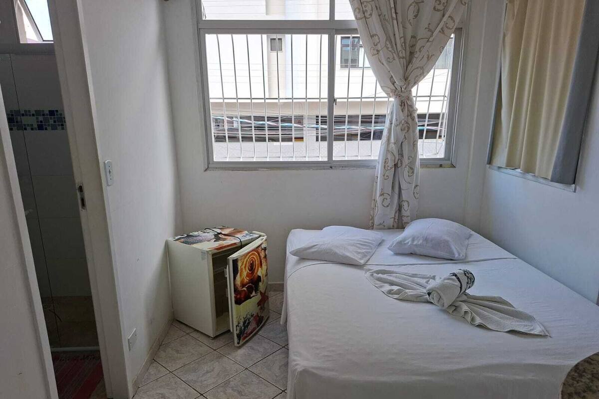 Quarto pequeno e limpo, com cama, janela com cortinas e uma mesinha com uma revista.