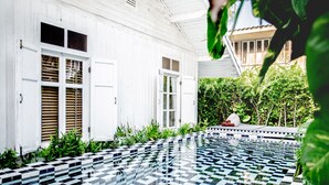 A pool with a checkered tile pattern, surrounded by lush greenery and a white building with open shutters. - The Hen Hua Hin Private Beach Villa (Hua Hin)