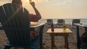 A beach scene with a person sitting on a blue chair, holding a drink, and facing the sunset. There are two tables with bottles and a glass on them. - Pacifico Paradise Condo, Playas del Coco, Guanacaste, Costa Rica (Coco)