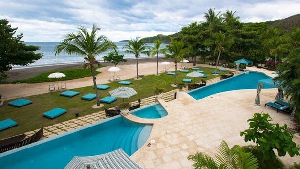 A pool area with lounge chairs and umbrellas, a beach in the background, and a view of the ocean. - Pacifico Paradise Condo, Playas del Coco, Guanacaste, Costa Rica (Coco)