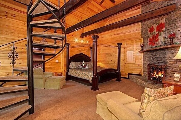 Interior de una cabaña de troncos con una escalera en espiral, una cama con dosel, una chimenea de piedra y un rincón de estar acogedor.