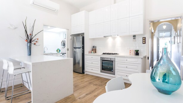 Una cocina moderna con armarios blancos, una isla central, electrodomésticos de acero inoxidable y una zona de comedor con sillas blancas.