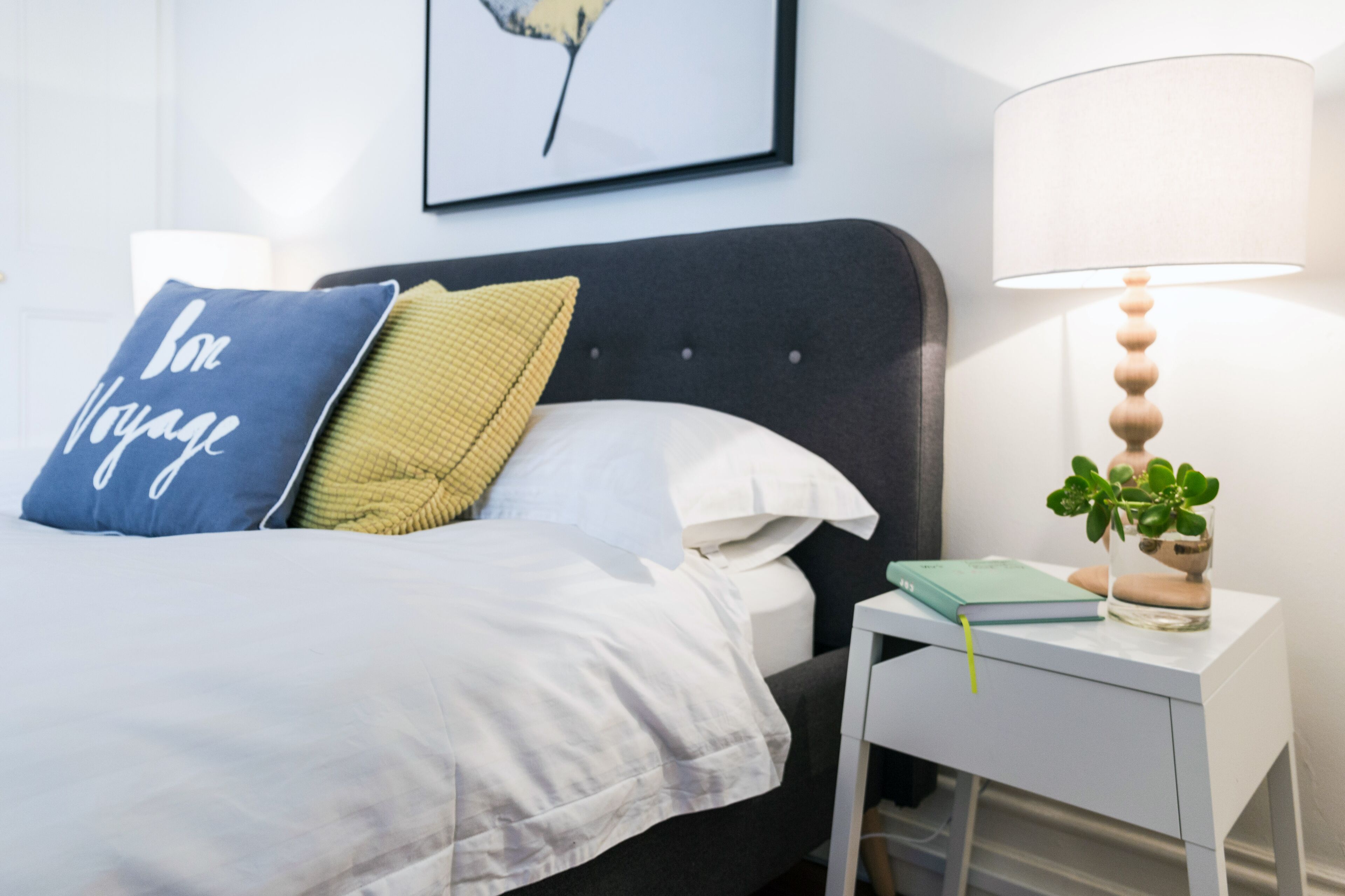 Una cama bien hecha con un almohadón azul, uno verde y uno blanco. Hay una mesita de noche con una lámpara, un cuaderno y una pequeña planta.