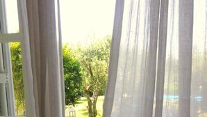 Une pièce dotée d’une grande fenêtre ornée de rideaux légers, d’une table blanche sur laquelle sont disposés divers objets, et d’où l’on peut admirer un arbre et la verdure environnante.