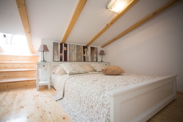 Une chambre avec un plafond en bois, un lit recouvert d’une literie à motifs, deux lampes de chevet et un grand tableau abstrait accroché au mur.