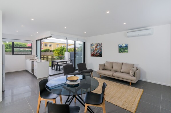 Una sala moderna con una mesa de comedor de vidrio, sillas negras, un sofá beige y un ventanal con vistas al exterior.