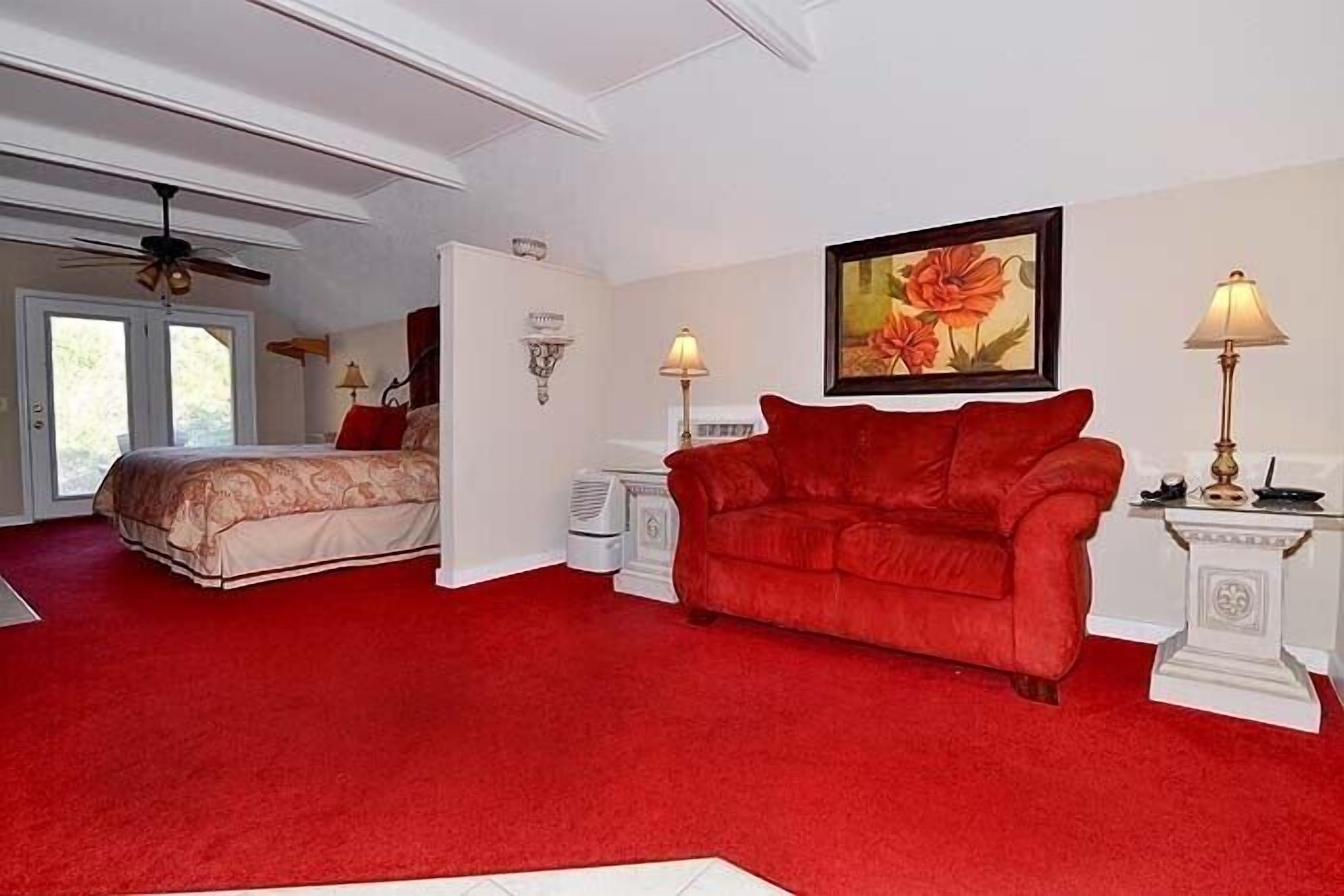 A room with a red carpet, a red sofa, a bed with a patterned bedspread, a ceiling fan, and a painting on the wall.