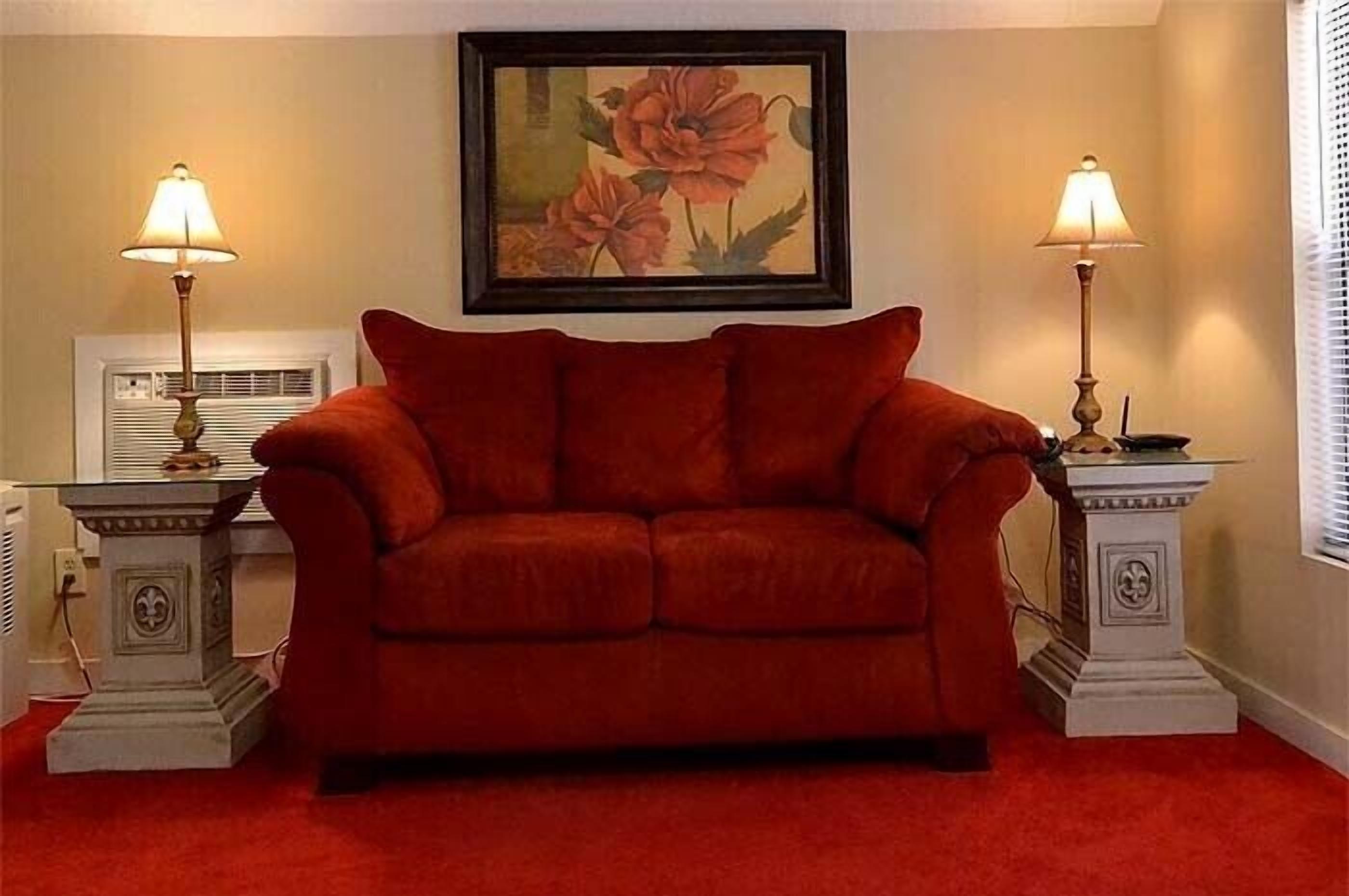 Un sofá rojo en una habitación con dos lámparas sobre mesitas auxiliares, un cuadro floral enmarcado en la pared y un radiador debajo del cuadro.