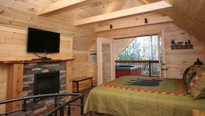 Une chambre située dans un chalet en rondins, avec une cheminée, un lit recouvert d’une courtepointe, un téléviseur fixé au mur et un balcon offrant une vue sur les arbres.