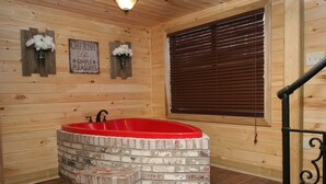 Une salle de bain rustique, avec une cabine en bois, une baignoire rouge, des murs en bois, une fenêtre avec des stores et une pancarte indiquant : « SAVOUREZ les PETITS BONHEURS de la vie ».