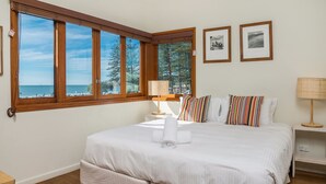 A bedroom with a large window overlooking the beach, a bed with white bedding, striped pillows, and a bedside table with a lamp.