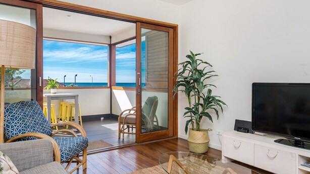 A modern living room with a large sliding glass door leading to a balcony with a view of the sea. There is a television, a plant, and a dining area with chairs and a table.