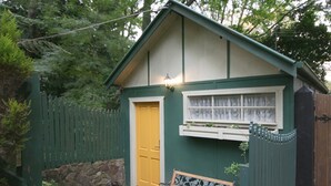 Un cobertizo pequeño de madera verde con una puerta amarilla, una ventana con cortinas blancas y una banca en el exterior.