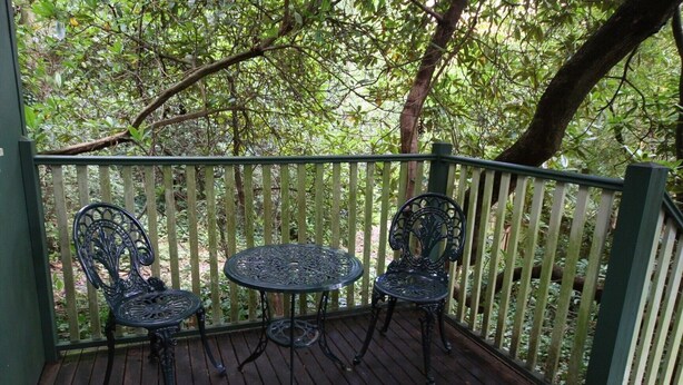 Una terraza de madera con una mesa de metal y dos sillas, rodeada por una valla de madera y árboles.