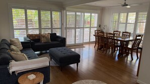 Ein geräumiges Wohnzimmer mit einem dunkelgrauen Sofa, passendem Hocker und einem Holzesstisch mit Stühlen. Große Fenster mit horizontalen Jalousien lassen natürliches Licht herein.