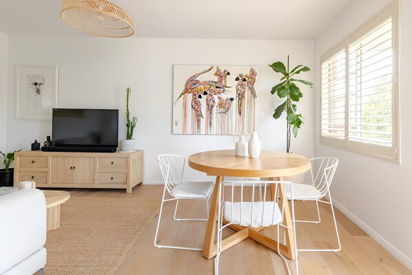 A modern living room with a round wooden dining table, white wire chairs, a flat-screen TV on a wooden cabinet, and a large abstract painting on the wall.