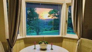 Una habitación con una mesa redonda, dos sillas, una ventana que ofrece vistas de un atardecer sobre un paisaje verde y una planta en maceta con velas sobre la mesa.