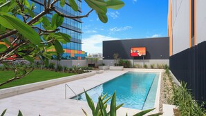 A modern swimming pool with a clear blue sky, surrounded by greenery and a modern building with large windows. - Executive 2BR Fortitude Valley apartment with pool and views (Fortitude Valley)