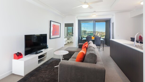 A modern living room with a flat-screen TV, a grey sofa with orange cushions, a small white table, and a ceiling fan. - Executive 2BR Fortitude Valley apartment with pool and views (Fortitude Valley)
