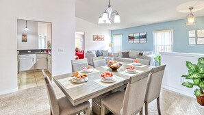 Uma sala de jantar com uma mesa de madeira posta para seis pessoas, rodeada por seis cadeiras. É visível uma cozinha ao fundo.