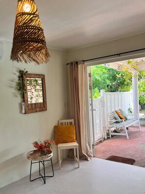 A room with a white chair, a small table with flowers, a woven light fixture, and a view of a garden through a window with curtains.