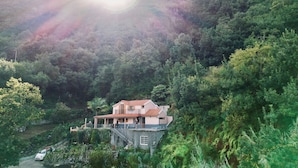 A two-story house with a balcony, surrounded by lush greenery and hills.