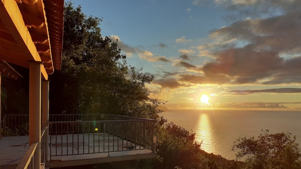A balcony overlooking a sunset over the sea, with a view of distant land and a bridge.
