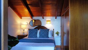 Un dormitorio con techo de madera, una cama con almohadas de diseño azulado, una lámpara de noche, un espejo y una planta pequeña sobre una mesita de noche.
