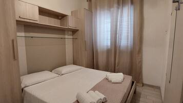 Un dormitorio pequeño y ordenado con una cama, dos almohadas, un estante sobre la cama y una ventana con cortinas.