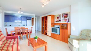 A modern living room with a sofa, dining table, chairs, a TV, and a kitchen area with cabinets and a microwave.