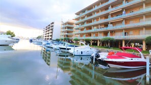 A marina with multiple boats docked, a multi-story building with balconies, and a clear sky.