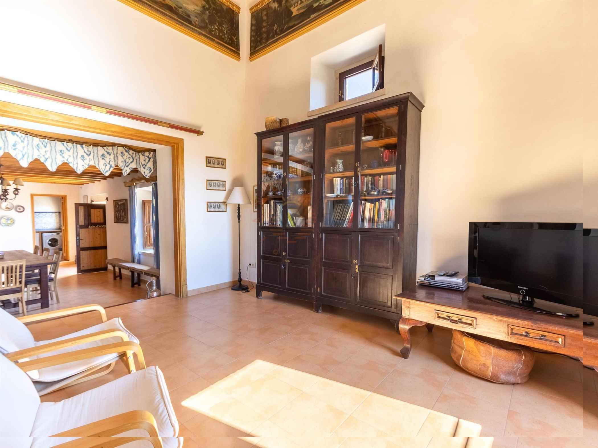 A spacious living room with a wooden coffee table, a dark wooden cabinet with glass doors, a flat-screen TV, and a dining area with wooden chairs and a bench.