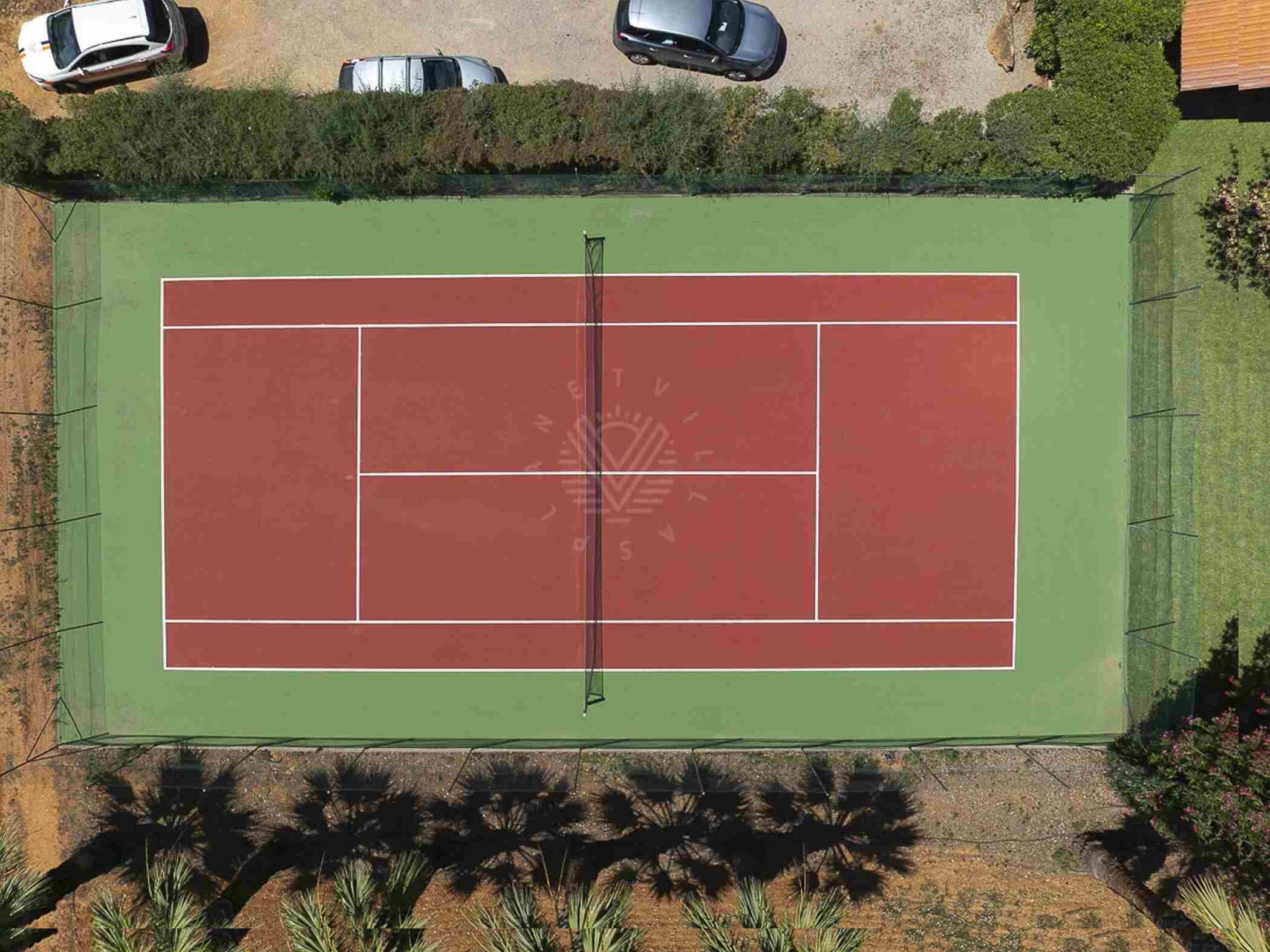 Vista aèria d'una pista de tennis envoltada de vegetació i cotxes estacionats.