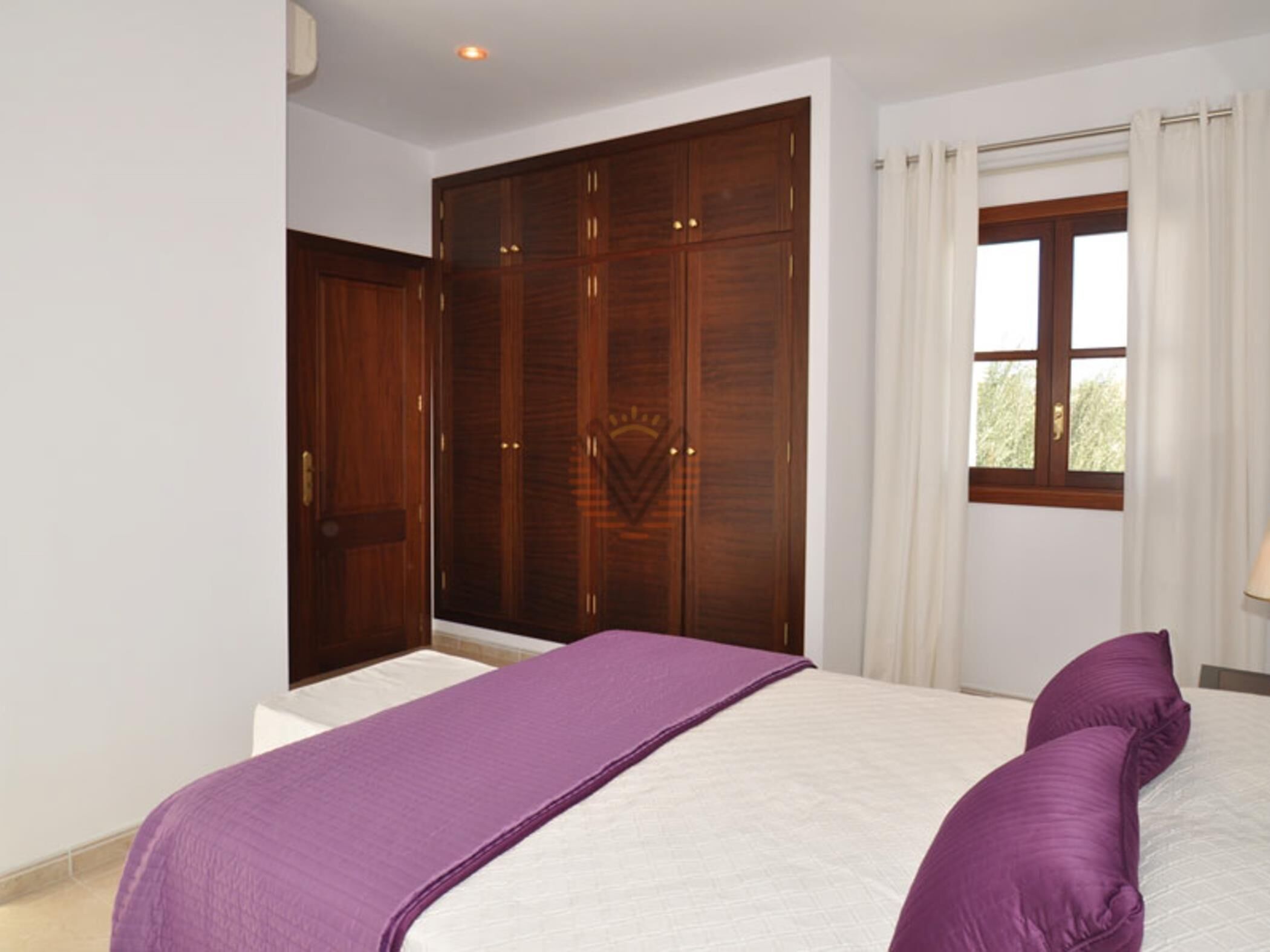 Een slaapkamer met een grote garderobe, een bed met een paars dekbed en een raam met gordijnen.