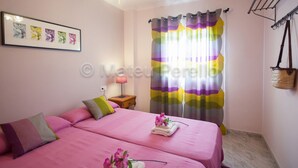 Une chambre avec deux lits, un ventilateur de plafond, une table de chevet, une lampe, une fenêtre avec des rideaux colorés et un porte-bagages fixé au mur.