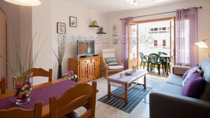 A living room with a sofa, a dining table with chairs, a TV on a wooden cabinet, and a balcony with chairs and plants. - Obelisc-1, 2 DB apartment near the beach (Can Picafort)