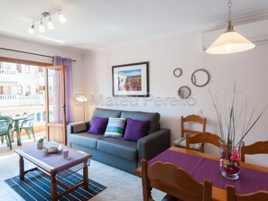 Ein modernes Wohnzimmer mit grauem Sofa, einem Holzesstisch und einem Esszimmerstuhl. An der Wand hängt ein gerahmtes Bild und auf dem Esstisch steht eine Vase mit Pflanzen.