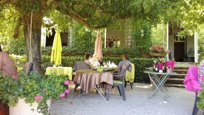 Un área de comedor al aire libre con mesas y sillas, rodeada de follaje y plantas en macetas.