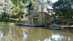 Una cabaña de madera con un porche y una pequeña terraza frente a un lago tranquilo, rodeada de árboles.