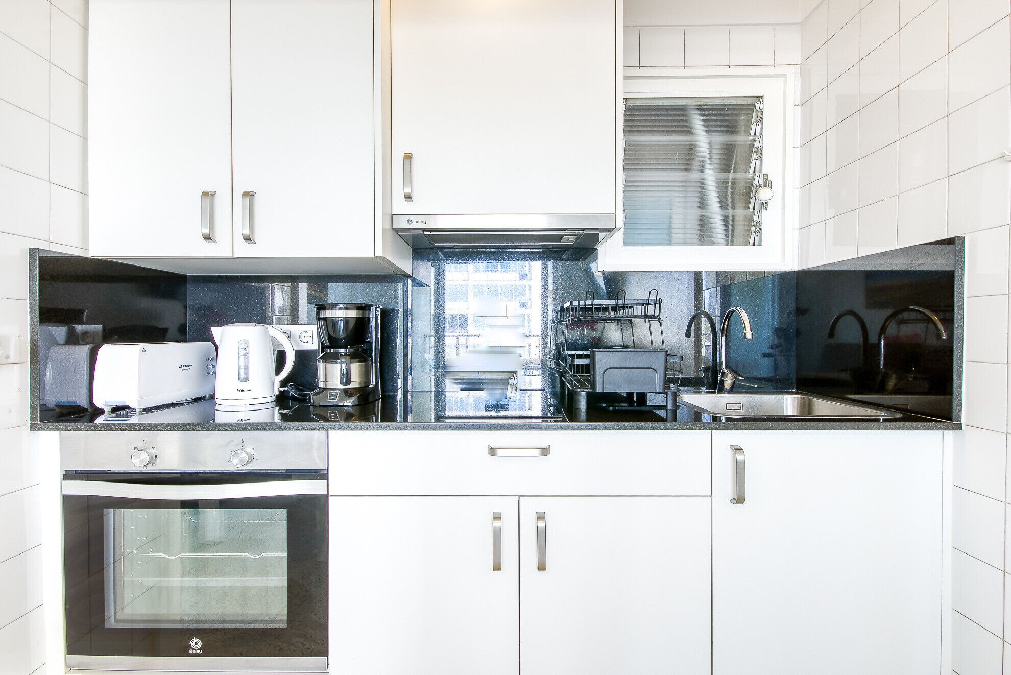 A modern kitchen with white cabinets, a coffee maker, a kettle, a dishwasher, and a built-in oven.