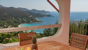 A wooden table with a fruit bowl and two wooden chairs on a balcony overlooking a coastal landscape.
