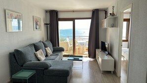 A modern living room with a grey sofa, a small table, a TV, and a balcony with a view of the sea. - Agi Panama 4C Apartamento for rent in Roses Girona. (Roses)