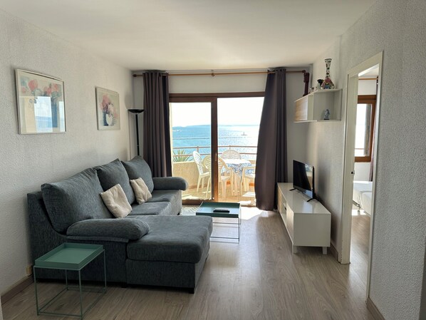 Een moderne woonkamer met een grijze bank, een klein tafeltje, een televisie en een balkon met uitzicht op zee.