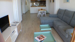 A modern living room with a grey sectional sofa, a small table with a magazine, a TV on a stand, and a kitchen in the background. - Agi Panama 4C Apartamento for rent in Roses Girona. (Roses)