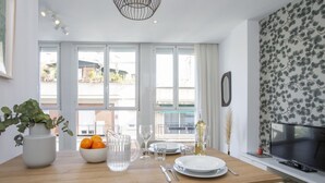 Un espace repas moderne avec une table en bois dressée pour quatre personnes, une plante en pot, un bol rempli d’oranges et un téléviseur posé sur un meuble.